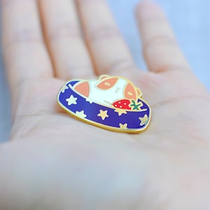 May include: Enamel pin featuring a sleeping animal with orange and white fur, nestled in a blue bed with gold stars. A red strawberry with green leaves rests beside the animal. The pin has a gold border.