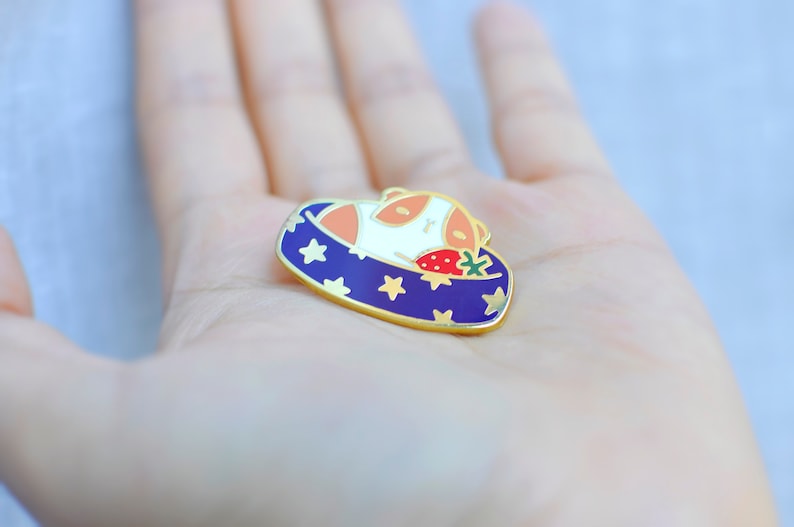 May include: Enamel pin featuring a sleeping animal with orange and white fur, nestled in a blue bed with gold stars. A red strawberry with green leaves rests beside the animal. The pin has a gold border.