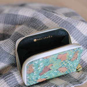Puede incluir: Cartera abierta con cremallera, con un estampado de criaturas marrones sobre un fondo turquesa. La cartera tiene un borde blanco y una cremallera dorada. El interior es verde oscuro con el texto dorado "noristudio".