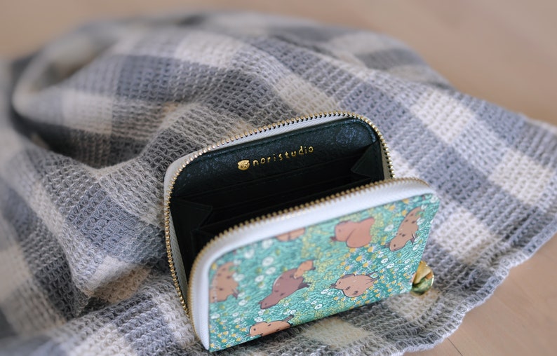 Puede incluir: Cartera abierta con cremallera, con un estampado de criaturas marrones sobre un fondo turquesa. La cartera tiene un borde blanco y una cremallera dorada. El interior es verde oscuro con el texto dorado "noristudio".