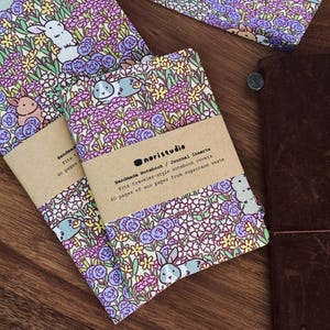 May include: Notebooks with floral and bunny pattern covers. The notebooks have a brown paper band with the text "Handmade Notebook / Journal Inserts". The covers feature a repeating pattern of bunnies, flowers, and foliage in shades of purple, yellow, and green.