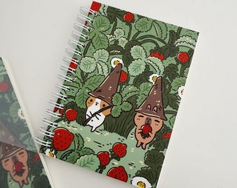Puede incluir: Un cuaderno con espiral con un diseño caprichoso que presenta personajes de dibujos animados con sombreros marrones en medio de un campo de fresas. La portada tiene un fondo verde con fresas rojas y flores blancas.