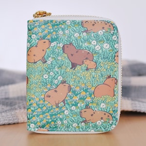 Puede incluir: Una cartera cuadrada con un estampado de carpinchos. La cartera presenta un dise&ntilde;o repetido de carpinchos marrones en un campo de flores amarillas y blancas sobre un fondo verde azulado. Tiene una cremallera dorada y un borde blanco.