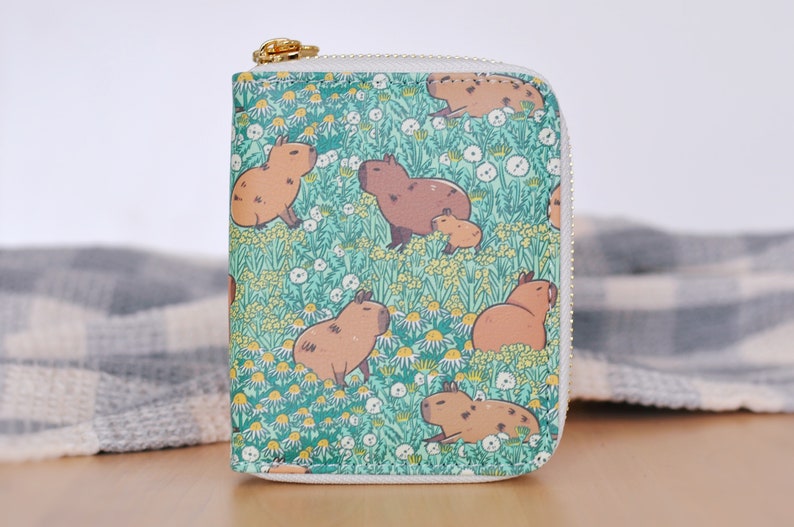 Puede incluir: Una cartera cuadrada con un estampado de carpinchos. La cartera presenta un dise&ntilde;o repetido de carpinchos marrones en un campo de flores amarillas y blancas sobre un fondo verde azulado. Tiene una cremallera dorada y un borde blanco.