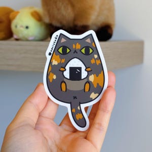 May include: A sticker featuring a cartoon cat holding a rice ball. The cat is gray with orange and tan spots, yellow eyes, and a black nose. The rice ball is square and black. The sticker has the text "noristudio".