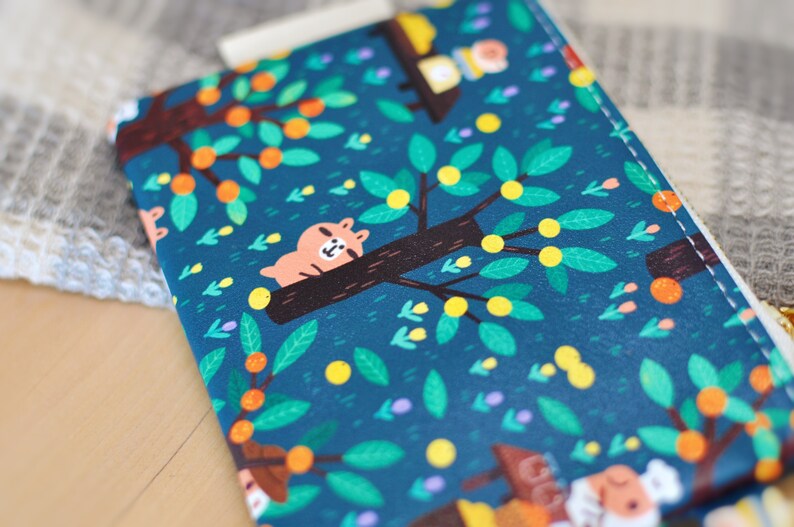 May include: A teal-colored zippered pouch with a whimsical tree and animal print. The design features a sleeping bear on a tree branch, orange fruits, and green leaves. The pouch has a white zipper and a gold-colored pull.