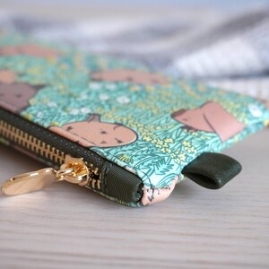 May include: A close-up of a zippered pouch with a whimsical pattern of light brown shapes on a teal background with yellow flowers. The zipper is gold, and the pull tab is dark green.