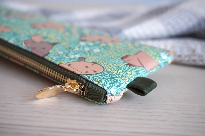 May include: A close-up of a zippered pouch with a whimsical pattern of light brown shapes on a teal background with yellow flowers. The zipper is gold, and the pull tab is dark green.