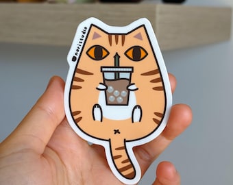 Könnte beinhalten: Ein Aufkleber mit einem orangefarbenen Cartoon-Katze mit braunen Streifen, die einen Bubble Tea hält. Der Bubble Tea befindet sich in einem transparenten Becher mit einem braunen Getränk und weißen Boba. Der Aufkleber trägt den Text "noristudio".