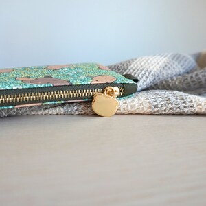 May include: A long, rectangular wallet with a teal background and a pattern of brown bear-like shapes. The wallet has a gold zipper and a gold bear-shaped zipper pull. A gray and white textured fabric is in the background.
