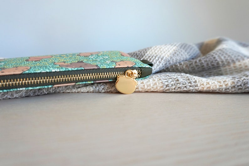 May include: A long, rectangular wallet with a teal background and a pattern of brown bear-like shapes. The wallet has a gold zipper and a gold bear-shaped zipper pull. A gray and white textured fabric is in the background.