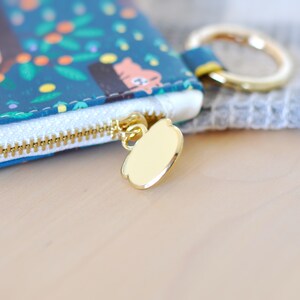 May include: Close-up of a teal zippered pouch with a gold-colored zipper and pull. The pouch features a whimsical pattern of trees, animals, and foliage. A gold ring is attached to the pouch. The background is blurred.