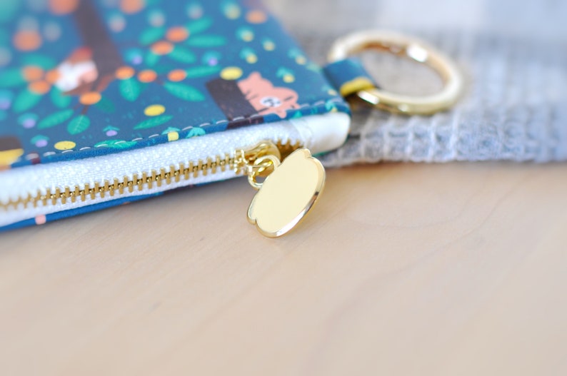May include: Close-up of a teal zippered pouch with a gold-colored zipper and pull. The pouch features a whimsical pattern of trees, animals, and foliage. A gold ring is attached to the pouch. The background is blurred.