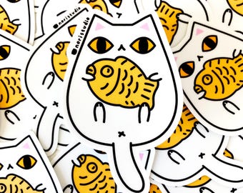 May include: White cat sticker with a yellow fish in its mouth. The cat has yellow eyes and pink ears. The sticker has a black outline and the text "noristudio" is visible on some stickers. The stickers are scattered.