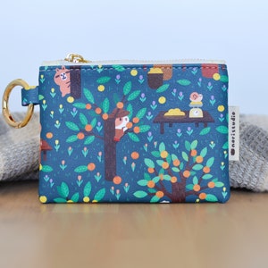 May include: A small, square coin purse with a whimsical illustration of a tree with oranges, animals, and a table. The purse has a gold zipper and a gold ring for attaching to a keychain. The background is a dark teal color.