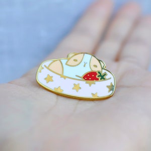 May include: A gold-colored enamel pin featuring a guinea pig in a bed with a strawberry. The bed is white with gold stars and a gold border. The guinea pig is light blue and tan.