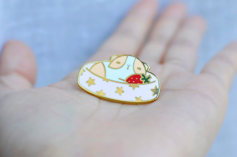 May include: A gold-colored enamel pin featuring a guinea pig in a bed with a strawberry. The bed is white with gold stars and a gold border. The guinea pig is light blue and tan.