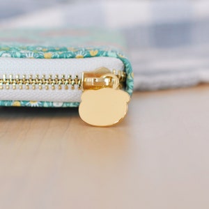 Puede incluir: Primer plano de una cartera con cremallera dorada y un tirador en forma de oso. La cartera tiene una cremallera blanca y una tela estampada en color turquesa y floral. La cremallera es dorada.