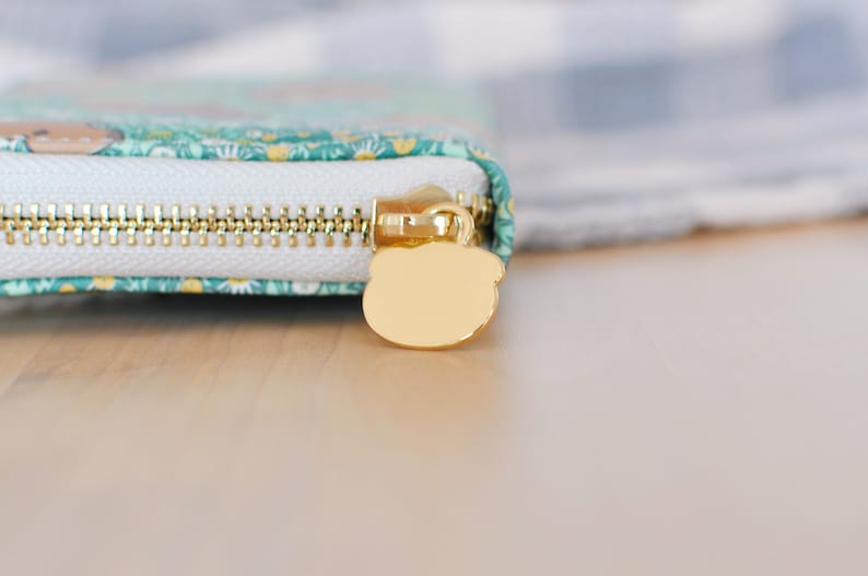 Puede incluir: Primer plano de una cartera con cremallera dorada y un tirador en forma de oso. La cartera tiene una cremallera blanca y una tela estampada en color turquesa y floral. La cremallera es dorada.