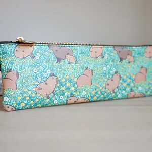 May include: A rectangular pencil case with a repeating pattern of capybaras in a field of flowers. The case has a teal background, a gold zipper, and a black leather pull tab. The capybaras are brown and tan.
