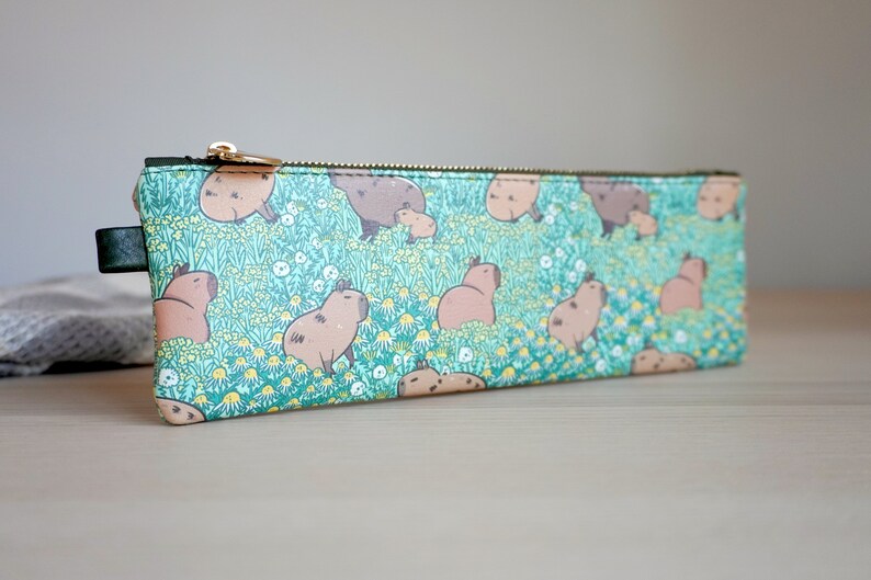 May include: A rectangular pencil case with a repeating pattern of capybaras in a field of flowers. The case has a teal background, a gold zipper, and a black leather pull tab. The capybaras are brown and tan.