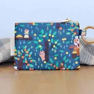 May include: A teal coin purse with a whimsical illustration of a tree with oranges, a squirrel, and a chef. The purse has a gold zipper and a gold ring for attaching keys. The design is printed on a rectangular pouch.