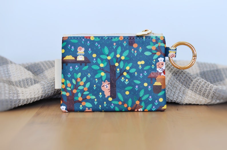 May include: A teal coin purse with a whimsical illustration of a tree with oranges, a squirrel, and a chef. The purse has a gold zipper and a gold ring for attaching keys. The design is printed on a rectangular pouch.
