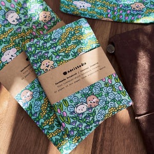 May include: Notebooks with a whimsical floral and animal pattern. The notebooks have a teal background with illustrations of flowers and cartoon animals. Each notebook is wrapped with a brown paper band that includes the text "noristudio".