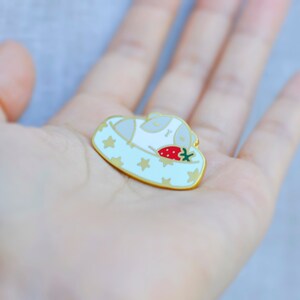 May include: A gold-colored enamel pin featuring a sleeping white animal in a light blue bed with gold stars. A red strawberry with green leaves rests beside the animal. The pin has a gold border.