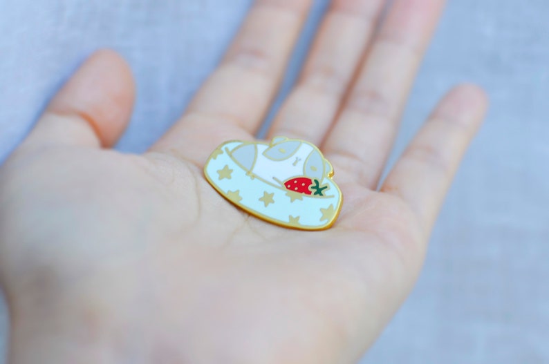 May include: A gold-colored enamel pin featuring a sleeping white animal in a light blue bed with gold stars. A red strawberry with green leaves rests beside the animal. The pin has a gold border.