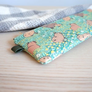 May include: A rectangular pencil case with a green background featuring a pattern of brown cartoon animals and white and yellow flowers. The case has a dark green leather loop attached to the side.