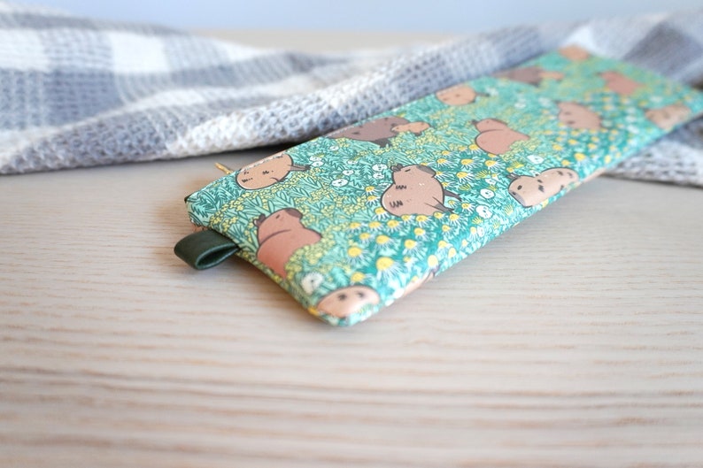 May include: A rectangular pencil case with a green background featuring a pattern of brown cartoon animals and white and yellow flowers. The case has a dark green leather loop attached to the side.