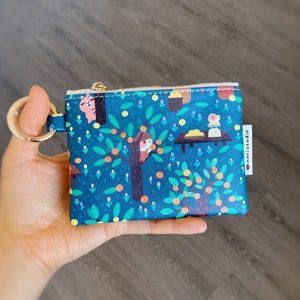 May include: A teal coin purse with a whimsical illustration of a tree with orange fruit, a squirrel, and a mouse. The purse has a gold zipper and a gold key ring. The brand name "noristudio" is printed on a white tag.