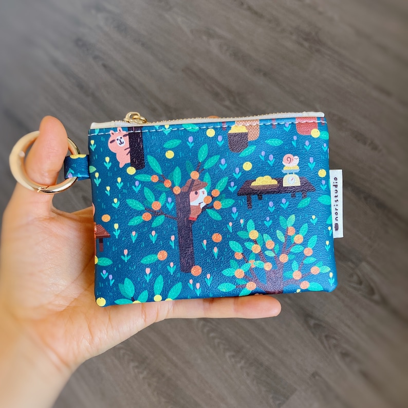 May include: A teal coin purse with a whimsical illustration of a tree with orange fruit, a squirrel, and a mouse. The purse has a gold zipper and a gold key ring. The brand name "noristudio" is printed on a white tag.