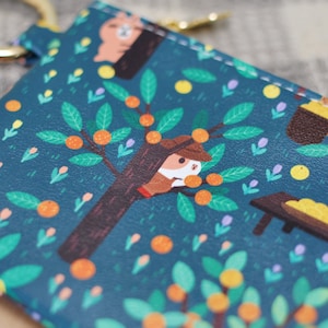May include: Teal-colored keychain wallet with a whimsical illustration of an orange grove. The design features a tree with oranges, a squirrel-like character, and a snail weighing fruit. A gold-colored ring is attached to the top.