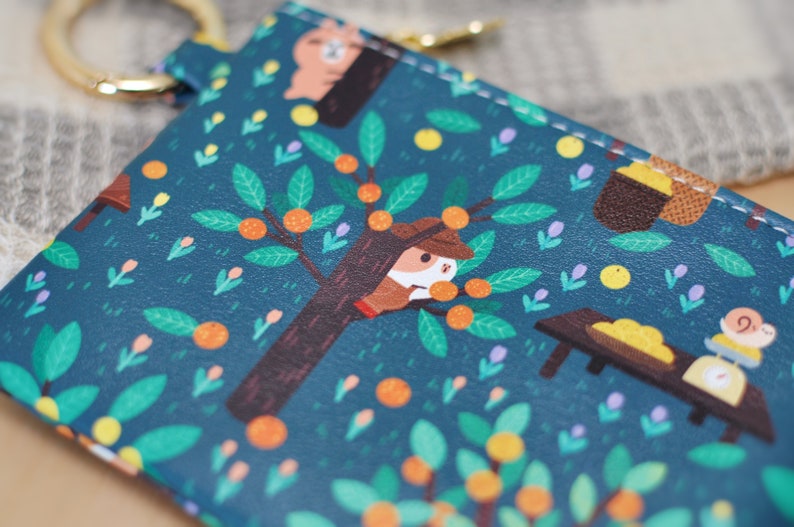 May include: Teal-colored keychain wallet with a whimsical illustration of an orange grove. The design features a tree with oranges, a squirrel-like character, and a snail weighing fruit. A gold-colored ring is attached to the top.