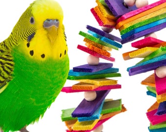 May include: A green and yellow parakeet bird perched on a white surface next to a colorful wooden bird toy. The toy is made of multiple brightly colored wooden pieces stacked on top of each other and connected by white beads.