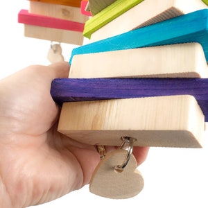 May include: A colorful wooden toy for birds, featuring a stack of brightly colored wooden blocks with a heart-shaped wooden charm hanging from the bottom. The blocks are painted in shades of pink, blue, green, purple, orange, and yellow.
