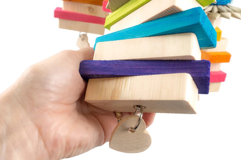 May include: A colorful wooden toy for birds, featuring a stack of brightly colored wooden blocks with a heart-shaped wooden charm hanging from the bottom. The blocks are painted in shades of pink, blue, green, purple, orange, and yellow.