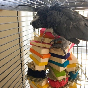 May include: A gray African grey parrot perched on a colorful wooden toy in a cage. The toy is made of stacked wooden blocks and has a variety of textures, including rope, straw, and fabric.
