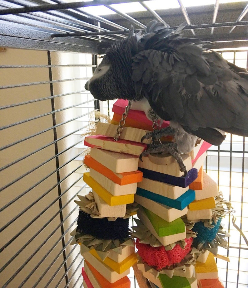 May include: A gray African grey parrot perched on a colorful wooden toy in a cage. The toy is made of stacked wooden blocks and has a variety of textures, including rope, straw, and fabric.