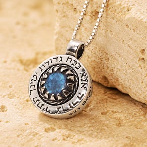 May include: A circular silver pendant necklace featuring a central blue gemstone surrounded by a textured metal border. The outer edge of the pendant is engraved with Hebrew text and the mark 925, suspended from a thin silver box chain.