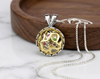 May include: A round pendant necklace featuring a silver outer frame and a gold-toned openwork cage design. The interior of the pendant contains small, multi-coloured gemstones including red, green, and purple stones. It hangs from a silver box chain.