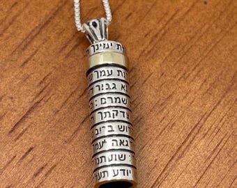 May include: A silver-toned pendant necklace with a cylindrical charm. The charm has Hebrew text engraved on it, with a gold-colored band near the top. The necklace is displayed on a wooden surface.