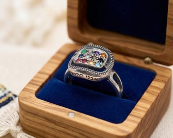 May include: A silver ring featuring a square face set with twelve small multicolored gemstones arranged in a grid. The band is engraved with Hebrew text along the side. The ring is displayed inside a small wooden box with a blue velvet interior.