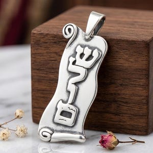 Puede incluir: Un colgante de plata con forma de pergamino con letras hebreas. El colgante tiene un lazo para una cadena y se muestra sobre un bloque de madera. Una pequeña rosa seca y una ramita de flores están en primer plano.