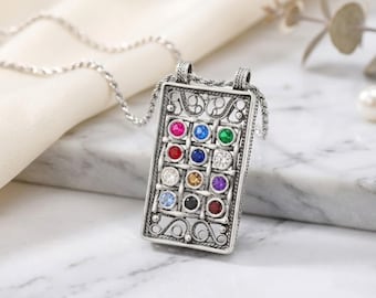 May include: A rectangular silver pendant necklace featuring an intricate openwork filigree frame. The center contains a grid of twelve small, round, multi-colored gemstones in shades of red, blue, green, white, yellow, purple, and black.