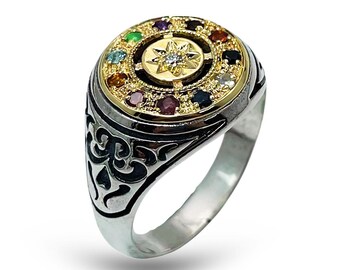 May include: A silver ring with a gold-colored circular top. The top features a central starburst design with a clear stone, surrounded by a ring of colorful gemstones. The ring's band has a detailed, dark pattern.