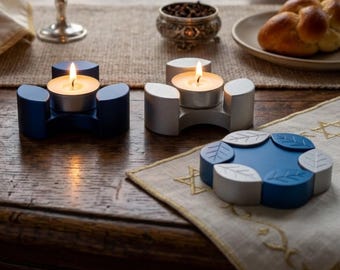 May include: Two tea light candle holders, one dark blue and one silver, each with a lit candle. A third circular object features a blue and silver leaf pattern. The holders have a three-pronged design, while the circular piece has six segments.
