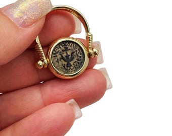 May include: A gold-colored ring featuring an ancient coin with a dark, detailed design. The coin is set in a circular bezel and attached to a curved band with decorative elements. The ring is held against a white background.